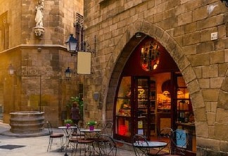 Construção antiga no Bairro Gótico em Barcelona. A construção remete ao perídodo medieval e em frente à ela estão mesas e cadeiras, presumindo que o lugar serve comidas.