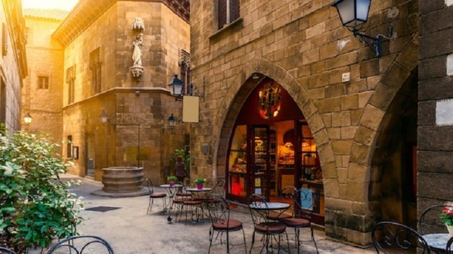 Construção antiga no Bairro Gótico em Barcelona. A construção remete ao perídodo medieval e em frente à ela estão mesas e cadeiras, presumindo que o lugar serve comidas.