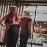 Dois manequins com roupas femininas de frente para a vitrine de uma loja. Os manequins vestem uma blusa vermelho escuro e calças escuras. Um deles está com a mão esquerda apoiada na cintura.
