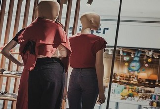 Dois manequins com roupas femininas de frente para a vitrine de uma loja. Os manequins vestem uma blusa vermelho escuro e calças escuras. Um deles está com a mão esquerda apoiada na cintura.