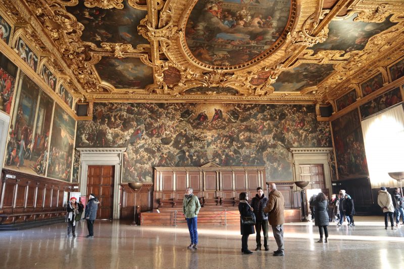 Interior do Palácio Ducal em Veneza. Os turistas observam as várias pinturas expostas nas paredes e no teto do lugar.