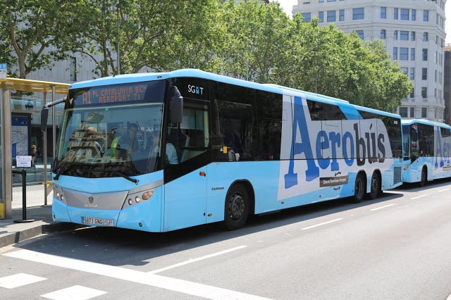 Ônibus Aerobús em Barcelona. O veículo é azul-claro e é possível notar um prédio ao fundo e árvores atrás do ônibus.