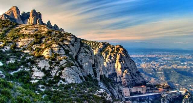 Mosteiro na montanha em Montserrat. Nota-se o céu azul com poucas nuvens. uma vegetação verde espalhada pelas pedras cinza da montanha e outras cidadezinhas ao longe, abaixo.