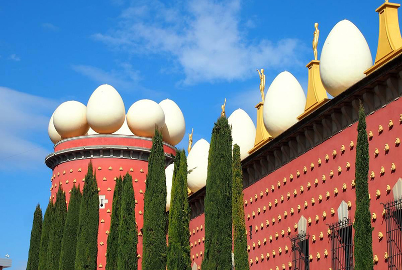 Fachada do Teatro-Museu Dalí em Figueres. Océu está azul e com poucas nuvens, a construção é cor de rosa e há pinheiros a sua frente.