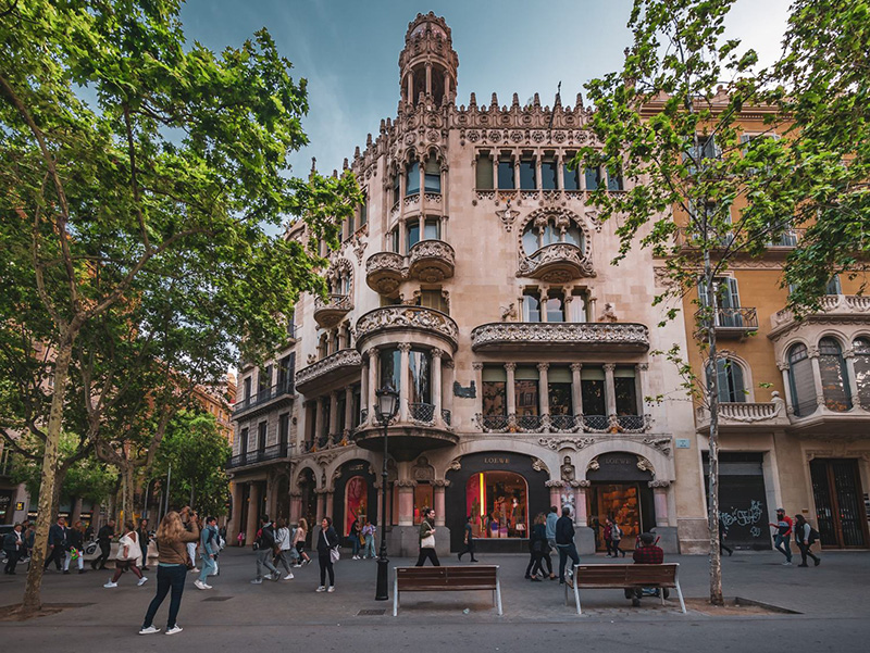 Vista de prédio em Passeig de Gràcia em Barcelona. Percebe-se pessoas transitando em frente à loja, duas árvores (cada uma ao lado) da loja e bancos do lado direito de um poste em frente ao prédio. 