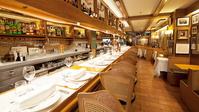 Interior do restaurante Botafumeiro em Barcelona. Nota-se um corredor composto por mesas e cadeiras, as mesas tem taças de vinho, pratos, talheres e guardanapos. Além disso, há uma espécie de bar para os atendentes servirem os clientes