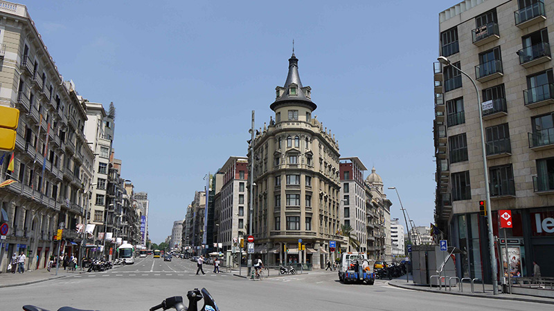 Vista de cruzamento na Rua Pelai em Barcelona. As ruas estão movimentadas com pessoas e carros e nota-se um prédio na esquina bem no meio da imagem