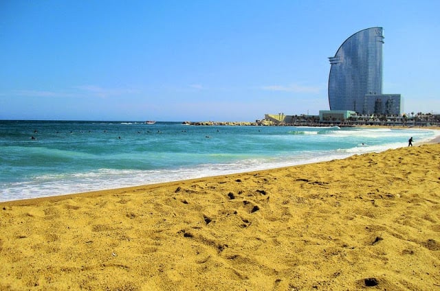 Vista da praia de Barceloneta em Barcelona. O céu está azul e a areia bem amarelada. Ao fundo, nota-se uma construção de um prédio ao fundo