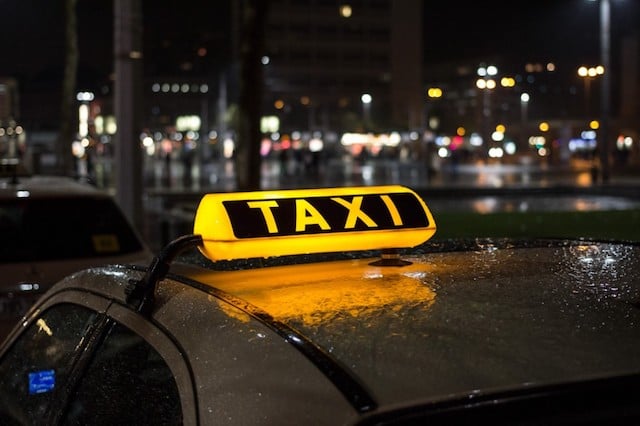 Carro andando pela rua de noite. A placa "Táxi" em cima do carro está acessa com a cor amarela bem forte