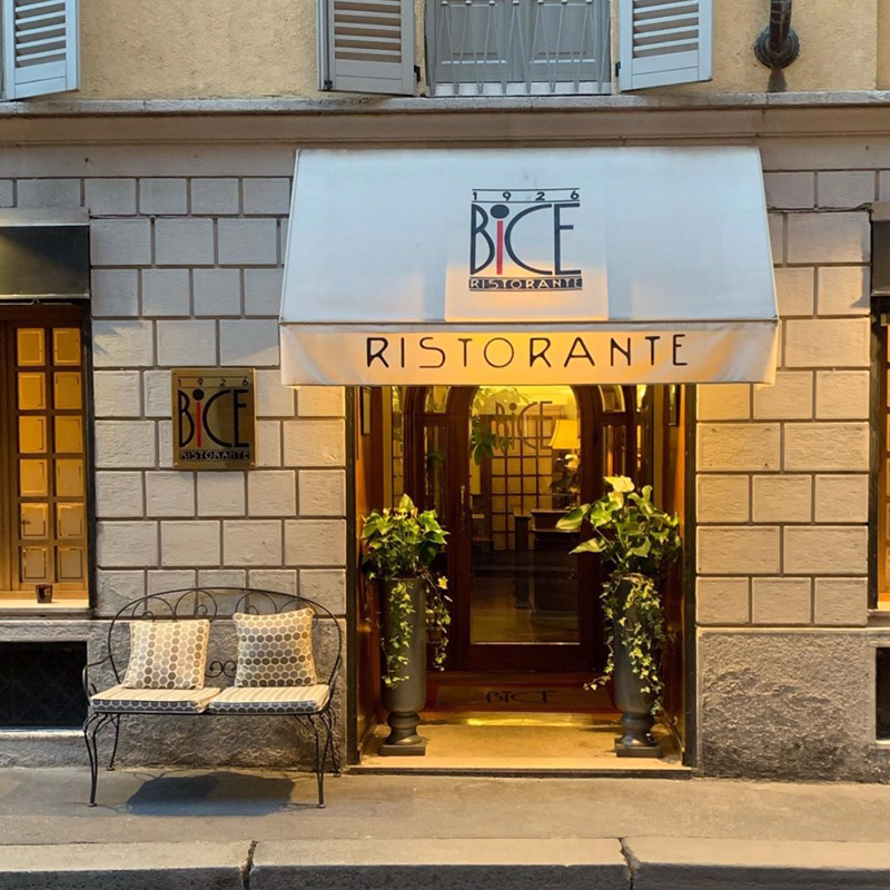 Fachada do restaurante Bice em Milão. Nota-se um assento antigo com duas almofadas ao lado da porta, além de dois vasos de plantas, um de cada lado, da porta de entrada.