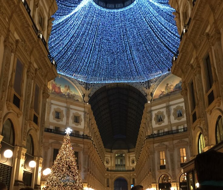 Interior da Galeria Vittorio Emanuele II em Milão. Nota-se a árvore de Natal totalmente iluminada e a cúpula da galeria enfeitada com pisca-piscas na cor azul. Tudo está muito iluminado