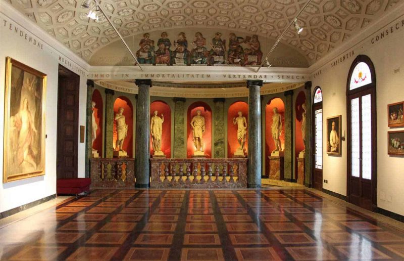 Interior da Pinacoteca Ambrosiana em Milão. Nota-se ao centro e mais ao fundo sete estátuas diferentes em um lugar à parte no museu. Perto do teto, acima das estátuas, está uma pintura de quatorze homens. Ainda, nas paredes ao redor, percebe-se que há quadros diversos. 