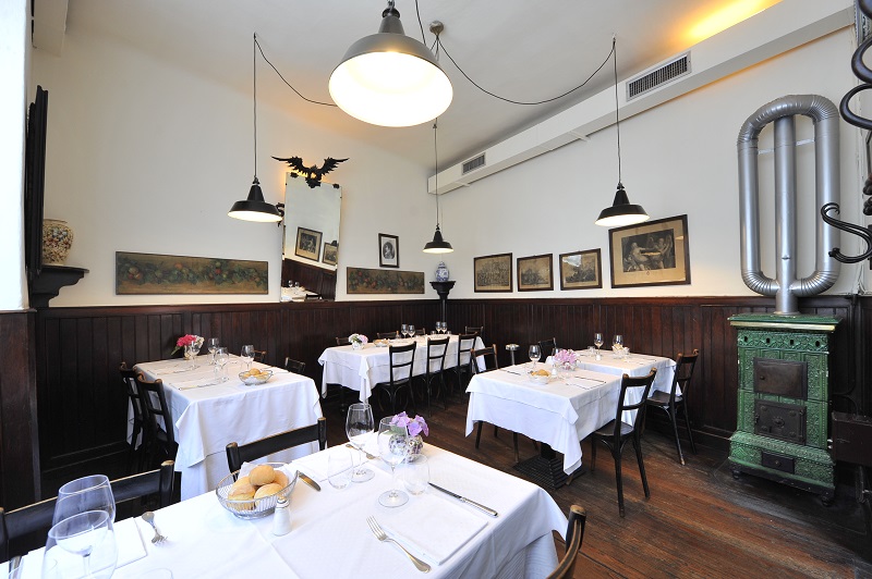 Interior do restaurante Antica Trattoria della Pesa em Milão. Notam-se mesas com toalhas brancas, móveis antigos, quadros nas paredes e lâmpadas.