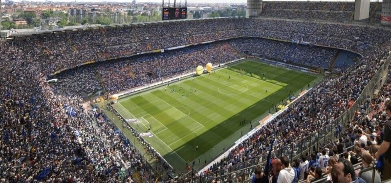 Vista do Estádio San Siro em Milão. O lugar está lotado e há jogadores em campo.