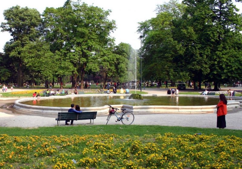 Vista do Parque Giardini Pubblici di Porta Venezia. Nota-se um jardim com flores amarelas em primeiro plano. Um casal sentado em um banco, uma bicicleta e uma pessoa em pé em segundo plano. E uma fonte com pessoas andando por perto e árvores ao fundo.