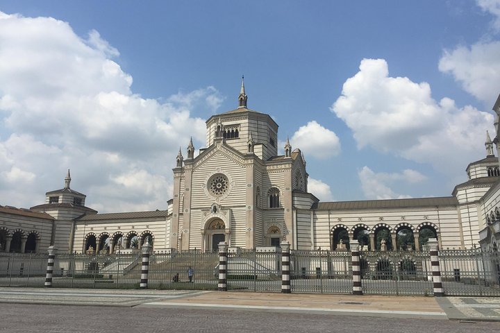 Fachada do Cemitério Monumental de Milão. O céu está com poucas nuvens e há uma espécie de capela na entrada. 