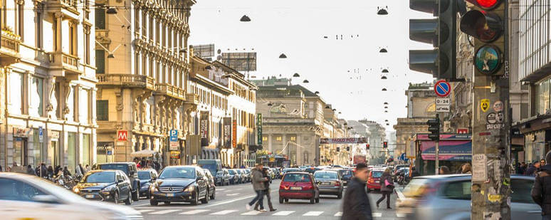 Vista da Avenida Corso Buenos Aires em Milão. Nota-se vários estabelecimentos, carros transitando na rua e pedestres andando sobre a faixa e pela avenida.