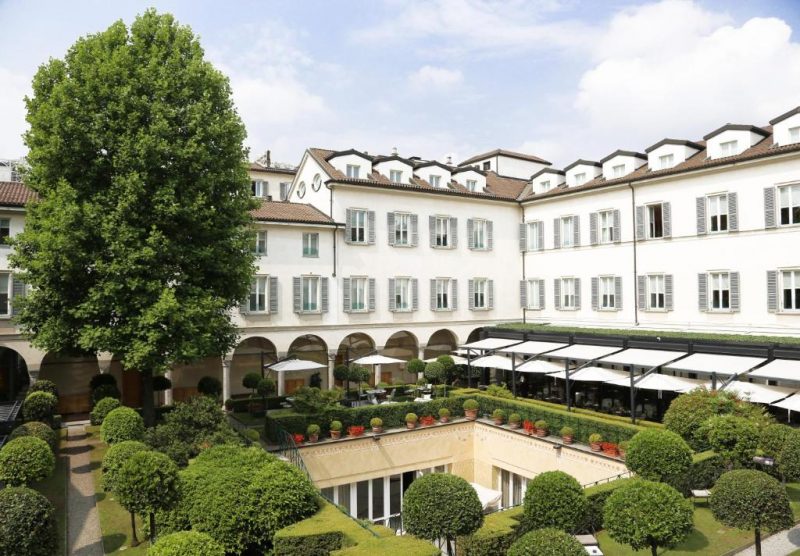 Vista do Hotel Four Seasons em Milão. Nota-se a estrutura do lugar parecida com uma casa antiga de cor branca. Além disso, percebe-se jardins com arbustos e uma grande árvore. 