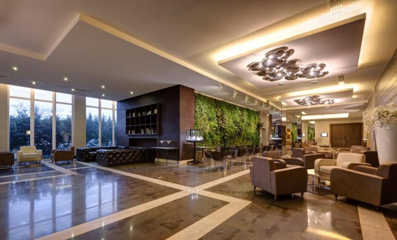 Interior do Klima Hotel Milano Fiere em Milão. O lugar é bem iluminado, possui móveis modernos e tons marrons e bege. Além disso, nota-se mais ao fundo, no centro da imagem, uma parede com plantas.