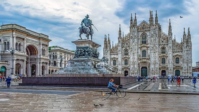 Vista do Monumento Equestre de Vittorio Emanuele II de frente para a Catedral de Milão. Do lado esquerdo, está a Galeria Vittorio Emanuele II. O céu está azul, com algumas nuvens e uma mulher anda de bicicleta à frente da estátua.