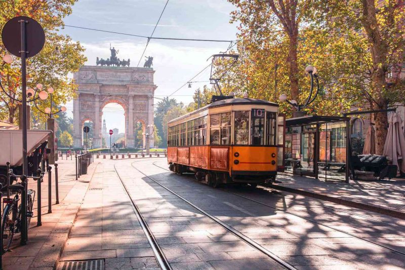 Vista do Arco della Pace em Milão. Ele está ao fundo e há um bondinho elétrico à frente, mais próximo ao primeiro plano da imagem. A cidade está pouco movimentada e está ensolarado.