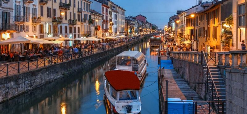 Região de Navigli de noite em Milão. Nota-se dois barcos passeando pelo rio no centro da imagem. Ao redor estão estabelecimentos com mesinhas do lado de fora e pessoas por lá.