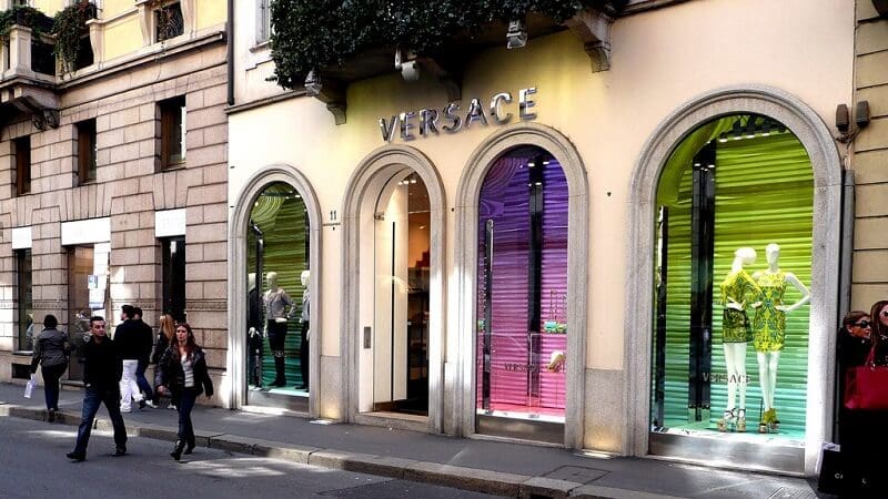 Fachada da loja Versace no quadrilátero da moda em Milão. Pessoas andam na rua e na calçada em frente ao lugar.