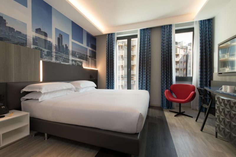 Quarto no iQ Hotel Milano. O lugar tem a decoração moderna com tons azuis e uma cadeira vermelha no canto direito.
