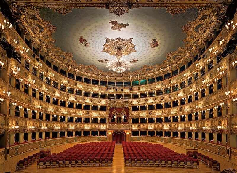 Interior da sala do Teatro La Fenice. O lugar tem a estrutura na cor dourada e o teto é azul com detalhes. Além do mais, o lugar está vazio e muito iluminado